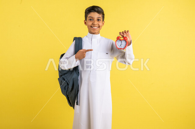 بورتريه لطالب عربي خليجي سعودي، يحمل الحقيبة المدرسية وبيده ساعة رقمية صغيرة، التذكيرات والتنبيهات لمواعيد المدرسة، يرتدي الزي السعودي التقليدي، الإستعداد للعام الدراسي الجديد، التعليم الدراسي في السعودية، العودة إلى المدارس، خلفية صفراء