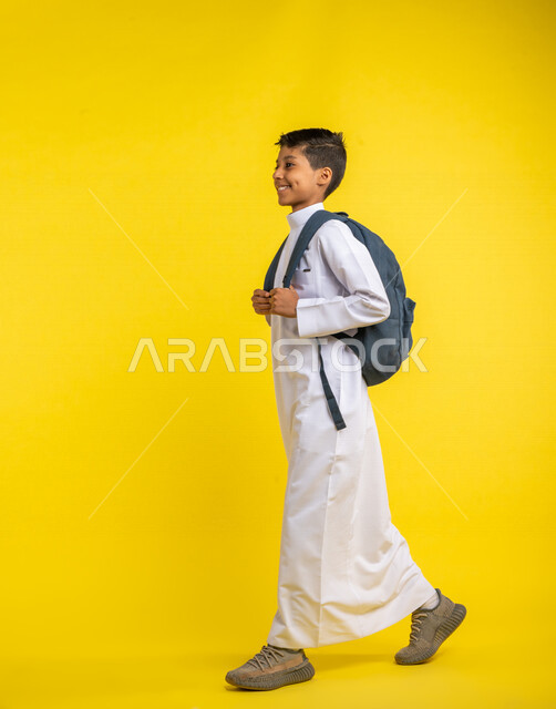 Portrait of a Saudi Arabian Gulf student, carrying a school bag, gestures to walk to school, wearing traditional Saudi dress, preparing for the new school year, school stage, school education in Saudi Arabia, yellow background, back to school