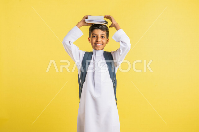 بورتريه لطالب عربي خليجي سعودي مبتسم ، يحمل الحقيبة المدرسية ، يضع الكتب المدرسية فوق رأسه ، يرتدي الزي السعودي التقليدي ، الإستعداد للعام الدراسي الجديد ، التعليم الدراسي في المملكة العربية السعودية ، خلفية صفراء ، العودة إلى المدارس