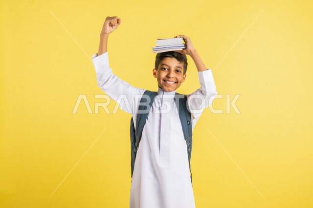 بورتريه لطالب عربي خليجي سعودي مبتسم ، يحمل الحقيبة المدرسية ، يضع الكتب المدرسية فوق رأسه ، يرتدي الزي السعودي التقليدي ، الإستعداد للعام الدراسي الجديد ، التعليم الدراسي في المملكة العربية السعودية ، خلفية صفراء ، العودة إلى المدارس