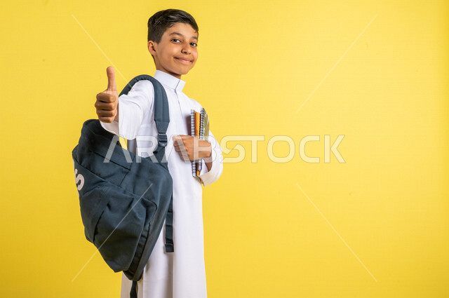 بورتريه لطالب عربي خليجي سعودي، يحمل الحقيبة المدرسية وبيده الكتب المدرسية، يشير بإصبعه الإبهام لأعلى بإيماءات تدل على الإعجاب، يرتدي الزي السعودي التقليدي، الإستعداد للعام الدراسي الجديد، التعليم الدراسي في السعودية، العودة إلى المدارس، خلفية صفراء