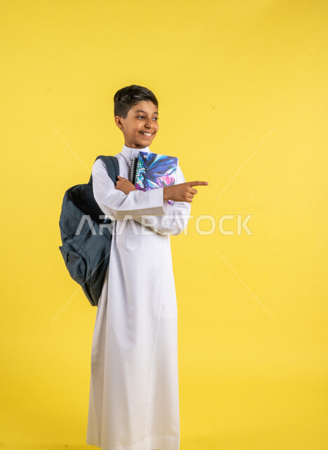 بورتريه لطالب عربي خليجي سعودي، يحمل الحقيبة المدرسية وبيده الكتب المدرسية، يشير بإصبعه السبابة لشيء ما ، يرتدي الزي السعودي التقليدي، الإستعداد للعام الدراسي الجديد، التعليم الدراسي في السعودية، العودة إلى المدارس، خلفية صفراء