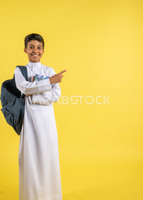 بورتريه لطالب عربي خليجي سعودي، يحمل الحقيبة المدرسية وبيده الكتب المدرسية، يشير بإصبعه السبابة لشيء ما ، يرتدي الزي السعودي التقليدي، الإستعداد للعام الدراسي الجديد، التعليم الدراسي في السعودية، العودة إلى المدارس، خلفية صفراء