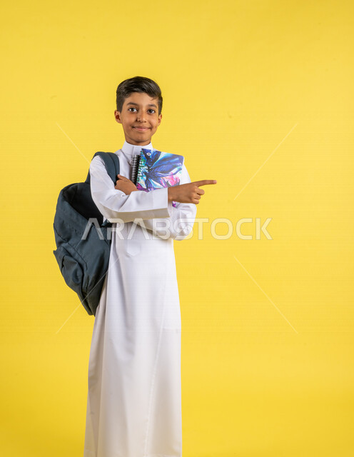 بورتريه لطالب عربي خليجي سعودي، يحمل الحقيبة المدرسية وبيده الكتب المدرسية، يشير بإصبعه السبابة لشيء ما ، يرتدي الزي السعودي التقليدي، الإستعداد للعام الدراسي الجديد، التعليم الدراسي في السعودية، العودة إلى المدارس، خلفية صفراء