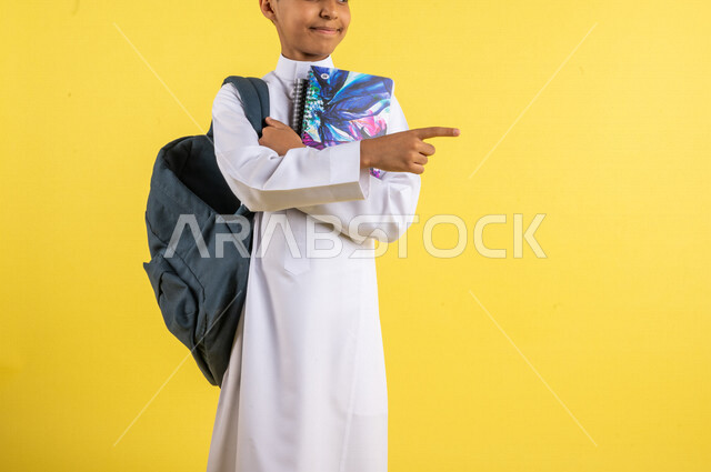 بورتريه لطالب عربي خليجي سعودي، يحمل الحقيبة المدرسية وبيده الكتب المدرسية، يشير بإصبعه السبابة لشيء ما ، يرتدي الزي السعودي التقليدي، الإستعداد للعام الدراسي الجديد، التعليم الدراسي في السعودية، العودة إلى المدارس، خلفية صفراء