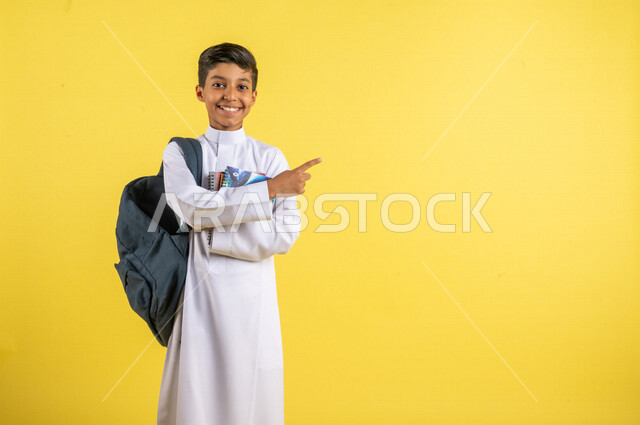 بورتريه لطالب عربي خليجي سعودي، يحمل الحقيبة المدرسية وبيده الكتب المدرسية، يشير بإصبعه السبابة لشيء ما ، يرتدي الزي السعودي التقليدي، الإستعداد للعام الدراسي الجديد، التعليم الدراسي في السعودية، العودة إلى المدارس، خلفية صفراء