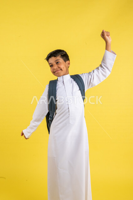 بورتريه لطالب عربي خليجي سعودي، يحمل الحقيبة المدرسية، يرفع يديه بإيماءات تدل على التمدد ، مفهوم النشاط والإستيقاظ الصباحي، يرتدي الزي السعودي التقليدي، الإستعداد للعام الدراسي الجديد، التعليم الدراسي في السعودية، العودة إلى المدارس، خلفية صفراء