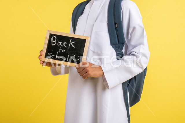 Portrait of a Saudi Arabian Gulf student, carrying a school bag, with a small blackboard written on it, back to school text, wearing traditional Saudi dress, preparing for the new school year, school education in Saudi Arabia, back to school, yellow backg