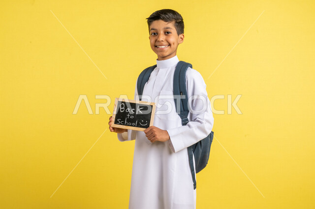بورتريه لطالب عربي خليجي سعودي، يحمل الحقيبة المدرسية، بيده سبورة صغيرة مكتوب عليها نص العودة إلى المدارس، يرتدي الزي السعودي التقليدي، الإستعداد للعام الدراسي الجديد، التعليم الدراسي في السعودية، العودة إلى المدارس، خلفية صفراء