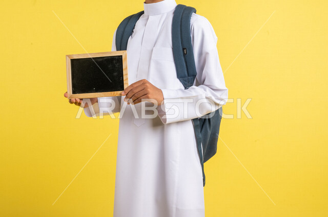 Portrait of a Saudi Arabian Gulf student carrying a school bag with a small empty blackboard in his hand, wearing traditional Saudi dress, preparing for the new school year, school education in Saudi Arabia, back to school, yellow background