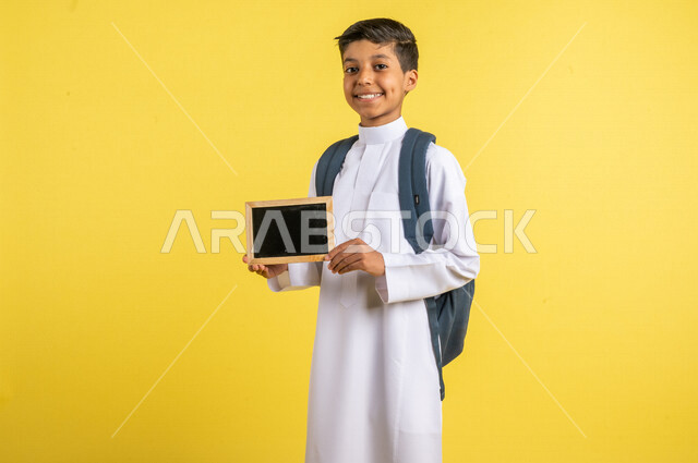 بورتريه لطالب عربي خليجي سعودي، يحمل الحقيبة المدرسية، بيده سبورة صغيرة فارغة، يرتدي الزي السعودي التقليدي، الإستعداد للعام الدراسي الجديد، التعليم الدراسي في السعودية، العودة إلى المدارس، خلفية صفراء