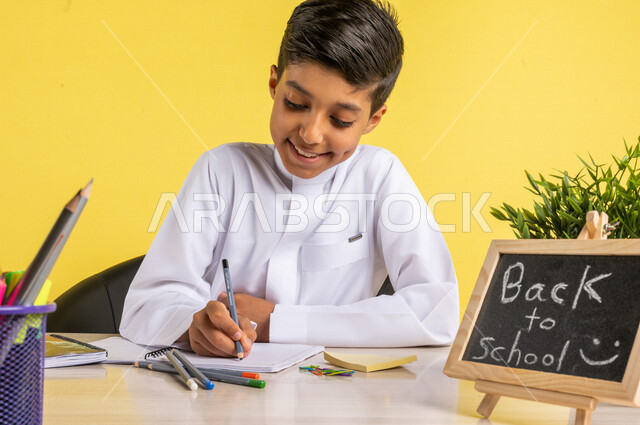 بورتريه لطالب عربي خليجي سعودي، يجلس على الطاولة، بجانبه مجموعة من الأقلام والكتب المدرسية، تأدية الواجبات المدرسية، سبورة صغيرة مكتوب عليها نص العودة إلى المدارس، الإستعداد للعام الدراسي الجديد، التعليم الدراسي في السعودية، العودة إلى المدارس،خلفية صفراء
