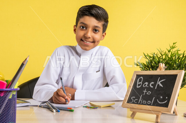 بورتريه لطالب عربي خليجي سعودي، يجلس على الطاولة، بجانبه مجموعة من الأقلام والكتب المدرسية، تأدية الواجبات المدرسية، سبورة صغيرة مكتوب عليها نص العودة إلى المدارس، الإستعداد للعام الدراسي الجديد، التعليم الدراسي في السعودية، العودة إلى المدارس،خلفية صفراء