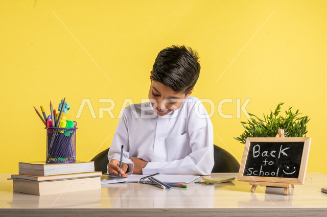 بورتريه لطالب عربي خليجي سعودي، يجلس على الطاولة، بجانبه مجموعة من الأقلام والكتب المدرسية، تأدية الواجبات المدرسية، سبورة صغيرة مكتوب عليها نص العودة إلى المدارس، الإستعداد للعام الدراسي الجديد، التعليم الدراسي في السعودية، العودة إلى المدارس،خلفية صفراء