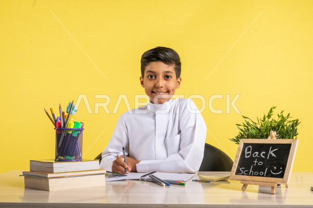 بورتريه لطالب عربي خليجي سعودي، يجلس على الطاولة، بجانبه مجموعة من الأقلام والكتب المدرسية، تأدية الواجبات المدرسية، سبورة صغيرة مكتوب عليها نص العودة إلى المدارس، الإستعداد للعام الدراسي الجديد، التعليم الدراسي في السعودية، العودة إلى المدارس،خلفية صفراء