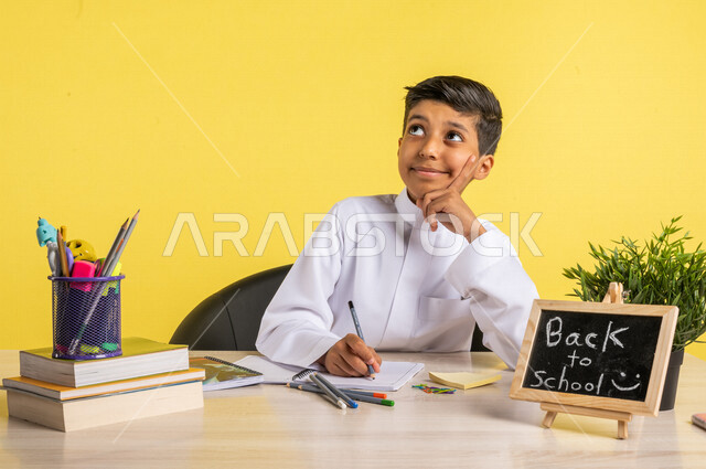 بورتريه لطالب عربي خليجي سعودي، يجلس على الطاولة، بجانبه مجموعة من الأقلام والكتب المدرسية، تأدية الواجبات المدرسية بإيماءات تدل على التفكير، سبورة صغيرة مكتوب عليها نص العودة إلى المدارس، الإستعداد للعام الدراسي الجديد، التعليم الدراسي في السعودية