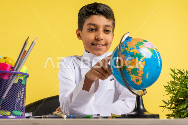 بورتريه لطالب عربي خليجي سعودي، يجلس على الطاولة، ينظر إلى مجسم الكرة الأرضية، بجانبه مجموعة من الأقلام والكتب المدرسية، مفهوم البحث والإستكشاف حول العالم، الإستعداد للعام الدراسي الجديد، التعليم الدراسي في السعودية، العودة إلى المدارس، خلفية صفراء
