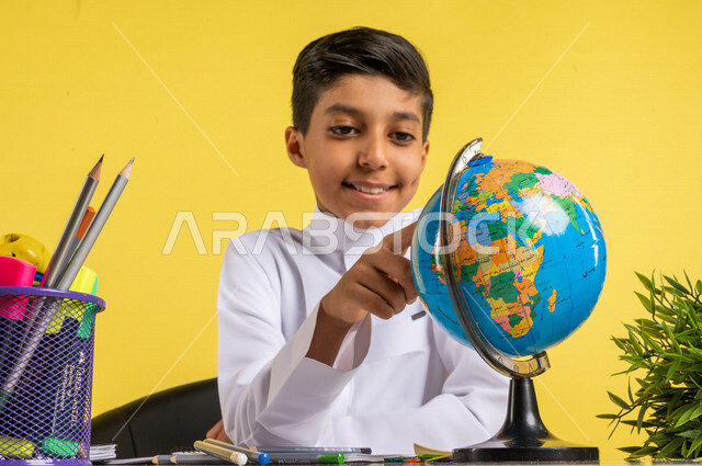 Portrait of a Saudi Arabian Gulf student, sitting at the table, looking at the globe, next to him a group of pens and textbooks, the concept of research and exploration around the world, preparing for the new school year, school education in Saudi Arabia,