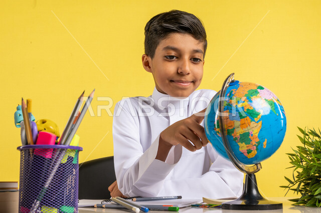 بورتريه لطالب عربي خليجي سعودي، يجلس على الطاولة، ينظر إلى مجسم الكرة الأرضية، بجانبه مجموعة من الأقلام والكتب المدرسية، مفهوم البحث والإستكشاف حول العالم، الإستعداد للعام الدراسي الجديد، التعليم الدراسي في السعودية، العودة إلى المدارس، خلفية صفراء