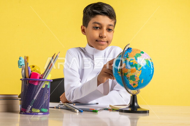 بورتريه لطالب عربي خليجي سعودي، يجلس على الطاولة، ينظر إلى مجسم الكرة الأرضية، بجانبه مجموعة من الأقلام والكتب المدرسية، مفهوم البحث والإستكشاف حول العالم، الإستعداد للعام الدراسي الجديد، التعليم الدراسي في السعودية، العودة إلى المدارس، خلفية صفراء