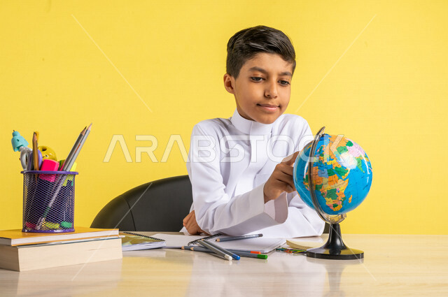 بورتريه لطالب عربي خليجي سعودي، يجلس على الطاولة، ينظر إلى مجسم الكرة الأرضية، بجانبه مجموعة من الأقلام والكتب المدرسية، مفهوم البحث والإستكشاف حول العالم، الإستعداد للعام الدراسي الجديد، التعليم الدراسي في السعودية، العودة إلى المدارس، خلفية صفراء