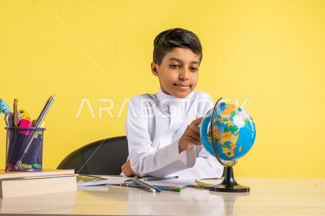 بورتريه لطالب عربي خليجي سعودي، يجلس على الطاولة، ينظر إلى مجسم الكرة الأرضية، بجانبه مجموعة من الأقلام والكتب المدرسية، مفهوم البحث والإستكشاف حول العالم، الإستعداد للعام الدراسي الجديد، التعليم الدراسي في السعودية، العودة إلى المدارس، خلفية صفراء