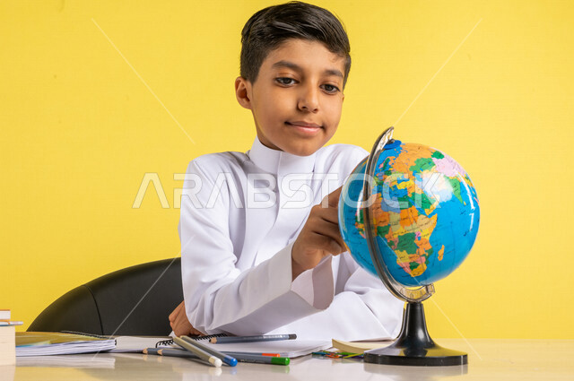 بورتريه لطالب عربي خليجي سعودي، يجلس على الطاولة، ينظر إلى مجسم الكرة الأرضية، بجانبه مجموعة من الأقلام والكتب المدرسية، مفهوم البحث والإستكشاف حول العالم، الإستعداد للعام الدراسي الجديد، التعليم الدراسي في السعودية، العودة إلى المدارس، خلفية صفراء