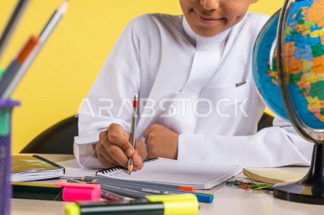 بورتريه لطالب عربي خليجي سعودي، بالزي السعودي التقليدي، يجلس على الطاولة، بجانبه مجموعة من الأقلام والكتب المدرسية ومجسم الكرة الأرضية، تأدية الواجبات المدرسية، الإستعداد للعام الدراسي الجديد، التعليم الدراسي في السعودية، العودة إلى المدارس، خلفية صفراء
