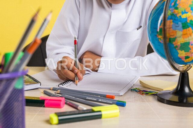 بورتريه لطالب عربي خليجي سعودي، بالزي السعودي التقليدي، يجلس على الطاولة، بجانبه مجموعة من الأقلام والكتب المدرسية ومجسم الكرة الأرضية، تأدية الواجبات المدرسية، الإستعداد للعام الدراسي الجديد، التعليم الدراسي في السعودية، العودة إلى المدارس، خلفية صفراء