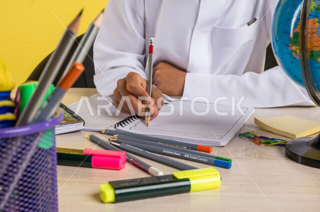 Portrait of a Saudi Arabian Gulf student, in traditional Saudi dress, sitting at a table, next to him, a group of pens, textbooks and a globe, doing homework, preparing for the new school year, school education in Saudi Arabia, back to school, yellow back