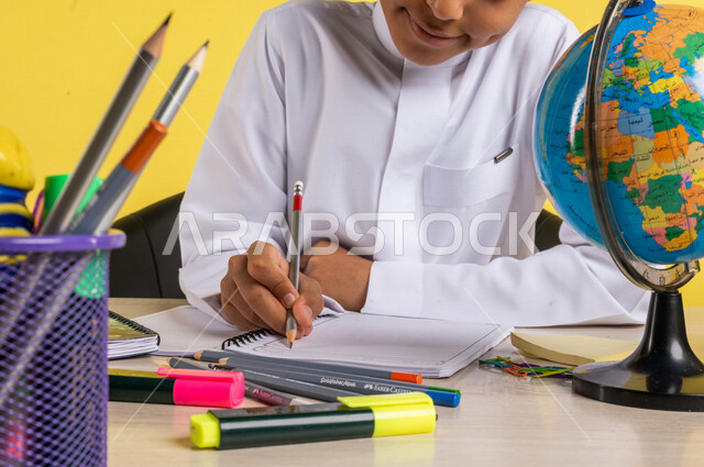 Portrait of a Saudi Arabian Gulf student, in traditional Saudi dress, sitting at a table, next to him, a group of pens, textbooks and a globe, doing homework, preparing for the new school year, school education in Saudi Arabia, back to school, yellow back