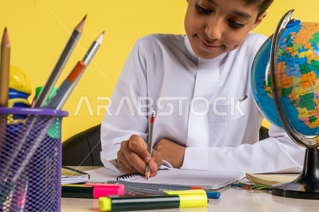 بورتريه لطالب عربي خليجي سعودي، بالزي السعودي التقليدي، يجلس على الطاولة، بجانبه مجموعة من الأقلام والكتب المدرسية ومجسم الكرة الأرضية، تأدية الواجبات المدرسية، الإستعداد للعام الدراسي الجديد، التعليم الدراسي في السعودية، العودة إلى المدارس، خلفية صفراء