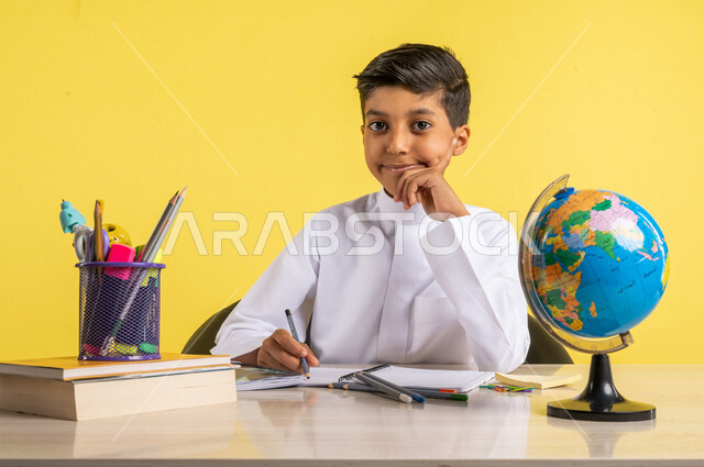 بورتريه لطالب عربي خليجي سعودي، بالزي السعودي التقليدي، يجلس على الطاولة، بجانبه مجموعة من الأقلام والكتب المدرسية ومجسم الكرة الأرضية، تأدية الواجبات المدرسية، الإستعداد للعام الدراسي الجديد، التعليم الدراسي في السعودية، العودة إلى المدارس، خلفية صفراء