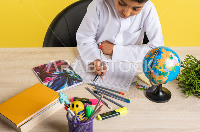 بورتريه لطالب عربي خليجي سعودي، بالزي السعودي التقليدي، يجلس على الطاولة، بجانبه مجموعة من الأقلام والكتب المدرسية ومجسم الكرة الأرضية، تأدية الواجبات المدرسية، الإستعداد للعام الدراسي الجديد، التعليم الدراسي في السعودية، العودة إلى المدارس، خلفية صفراء