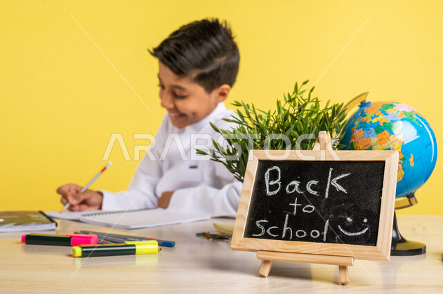 Portrait of a Saudi Arabian Gulf student, sitting at the table, next to him is a group of pens, textbooks and a globe, doing homework, a small blackboard with the text back to school written on it, preparing for the new school year, school education in Sa