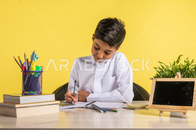 بورتريه لطالب عربي خليجي سعودي، بالزي السعودي التقليدي، يجلس على الطاولة، بجانبه مجموعة من الأقلام والكتب المدرسية، تأدية الواجبات المدرسية، سبورة صغيرة فارغة، الإستعداد للعام الدراسي الجديد، التعليم الدراسي في السعودية، العودة إلى المدارس، خلفية صفراء