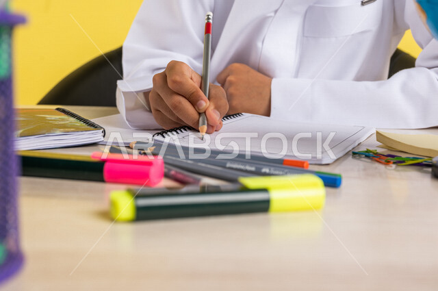 بورتريه بصورة مقربة لطالب عربي خليجي سعودي، يرتدي الزي السعودي التقليدي، يجلس على الطاولة، بجانبه مجموعة من الأقلام والكتب المدرسية، تأدية الواجبات المدرسية، الإستعداد للعام الدراسي الجديد، التعليم الدراسي في السعودية، العودة إلى المدارس، خلفية صفراء