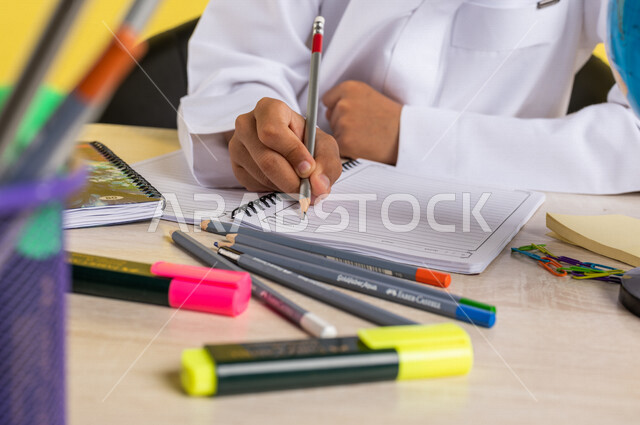 بورتريه بصورة مقربة لطالب عربي خليجي سعودي، يرتدي الزي السعودي التقليدي، يجلس على الطاولة، بجانبه مجموعة من الأقلام والكتب المدرسية، تأدية الواجبات المدرسية، الإستعداد للعام الدراسي الجديد، التعليم الدراسي في السعودية، العودة إلى المدارس، خلفية صفراء