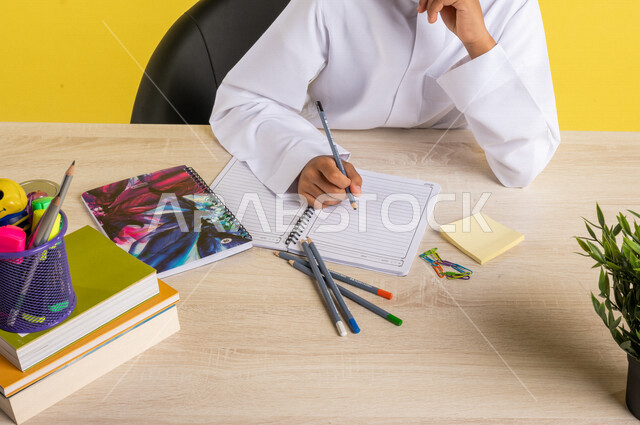 بورتريه بصورة مقربة لطالب عربي خليجي سعودي، يرتدي الزي السعودي التقليدي، يجلس على الطاولة، بجانبه مجموعة من الأقلام والكتب المدرسية، تأدية الواجبات المدرسية، الإستعداد للعام الدراسي الجديد، التعليم الدراسي في السعودية، العودة إلى المدارس، خلفية صفراء