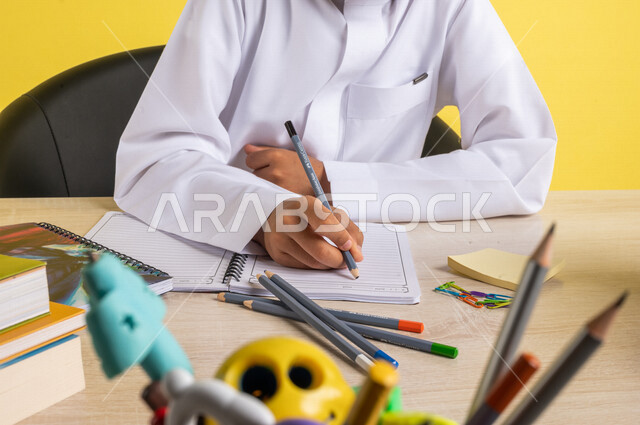 بورتريه بصورة مقربة لطالب عربي خليجي سعودي، يرتدي الزي السعودي التقليدي، يجلس على الطاولة، بجانبه مجموعة من الأقلام والكتب المدرسية، تأدية الواجبات المدرسية، الإستعداد للعام الدراسي الجديد، التعليم الدراسي في السعودية، العودة إلى المدارس، خلفية صفراء