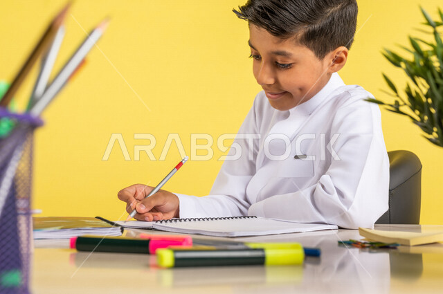 بورتريه لطالب عربي خليجي سعودي، يرتدي الزي السعودي التقليدي، يجلس على الطاولة، بجانبه مجموعة من الأقلام والكتب المدرسية، تأدية الواجبات المدرسية، الإستعداد للعام الدراسي الجديد، التعليم الدراسي في السعودية، العودة إلى المدارس، خلفية صفراء