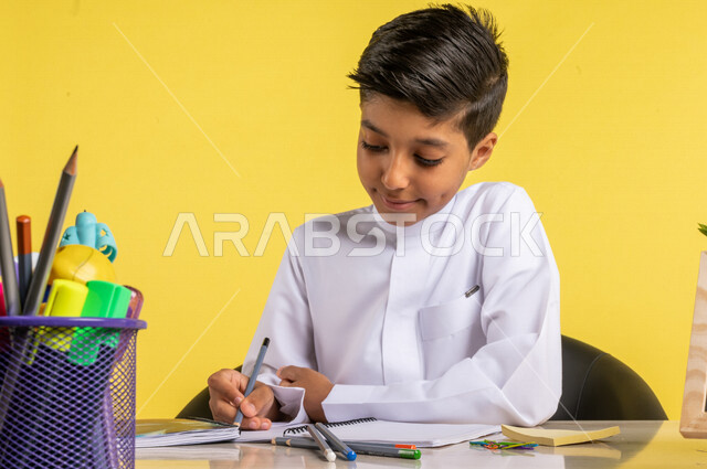 بورتريه لطالب عربي خليجي سعودي، يرتدي الزي السعودي التقليدي، يجلس على الطاولة، بجانبه مجموعة من الأقلام والكتب المدرسية، تأدية الواجبات المدرسية، الإستعداد للعام الدراسي الجديد، التعليم الدراسي في السعودية، العودة إلى المدارس، خلفية صفراء