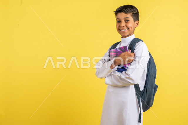 بورتريه لطالب عربي خليجي سعودي، يرتدي الزي السعودي التقليدي، يحمل الحقيبة المدرسية وبيده الكتب المدرسية، الإستعداد للعام الدراسي الجديد، التعليم الدراسي في المملكة العربية السعودية، العودة إلى المدارس، خلفية صفراء