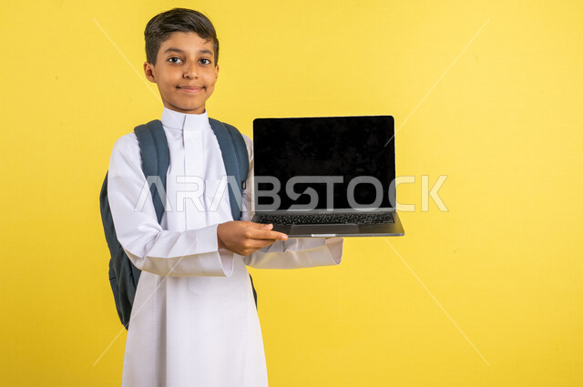 Portrait of a Saudi Arabian Gulf student, carrying a school backpack and a laptop in his hand, distance education, e-learning, following up on lessons and assignments online, preparing for the new school year, school education in Saudi Arabia, yellow back