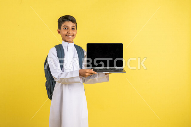 Portrait of a Saudi Arabian Gulf student, carrying a school backpack and a laptop in his hand, distance education, e-learning, following up on lessons and assignments online, preparing for the new school year, school education in Saudi Arabia, yellow back