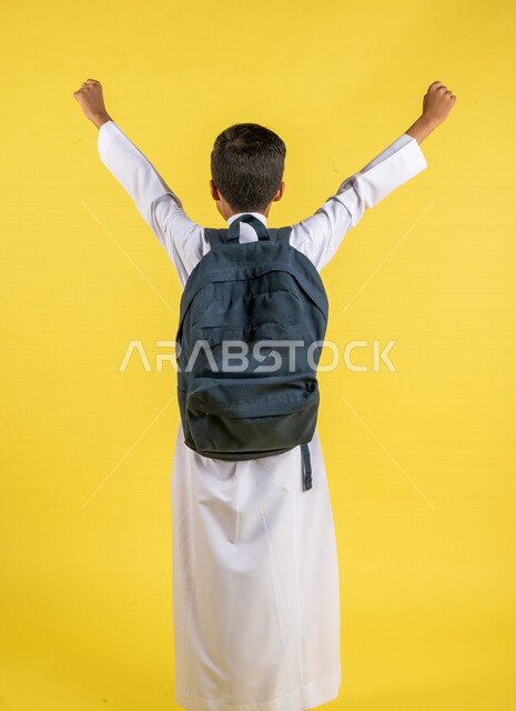 بورتريه بزاوية من الخلف لطالب عربي خليجي سعودي، بالزي السعودي التقليدي، يحمل حقيبة الظهر المدرسية، يرفع يديه لأعلى بإيماءات تدل على الحماس، الإستعداد للعام الدراسي الجديد، التعليم الدراسي في السعودية، خلفية صفراء، العودة إلى المدارس