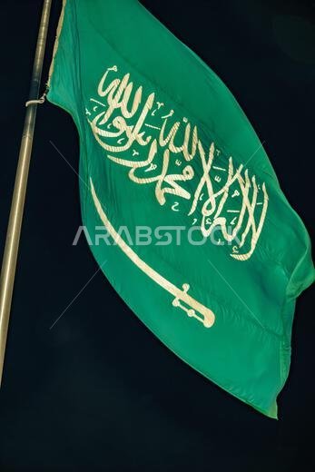 صورة مقرّبة لعلم المملكة العربية السعودية معلّق على عمود مرتفع. وهو مصنوع من قماش أخضر اللون وعليه كتابة بيضاء.