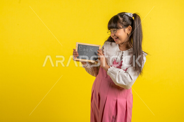 Portrait of a Saudi Arabian Gulf student, wearing a school uniform, holding a small blackboard with the text back to school written on it, preparing for the new school year, school education in Saudi Arabia, back to school, yellow background