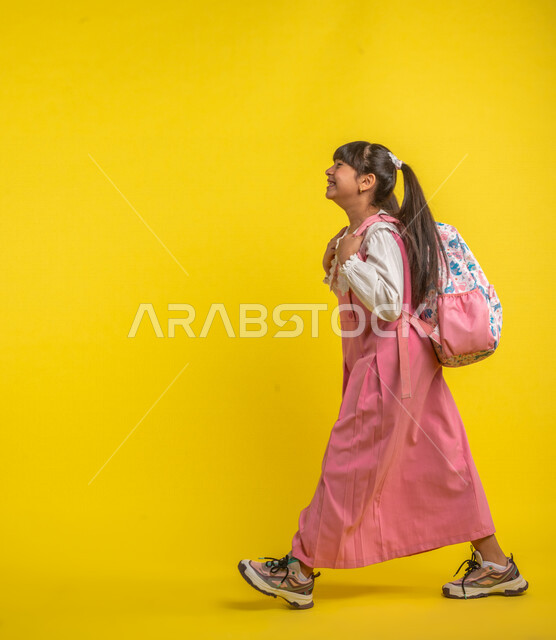 بورتريه لطالبة عربية خليجية سعودية، ترتدي الزي المدرسي، تحمل الحقيبة المدرسية، إيماءات تدل على السعادة ، السير والتوجه إلى المدرسة، الإستعداد للعام الدراسي الجديد، مرحلة دراسية، التعليم الدراسي في السعودية، خلفية صفراء، العودة إلى المدارس