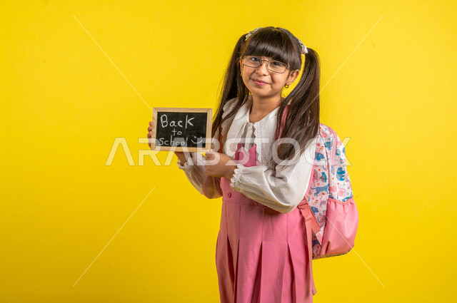 Portrait of a Saudi Arabian Gulf student, wearing a school uniform, carrying a school bag and a small blackboard with the text back to school written on it, preparing for the new school year, school education in Saudi Arabia, back to school, yellow backgr
